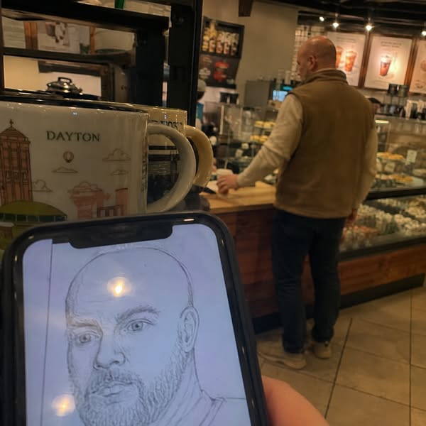 In a coffee shop, a man stands at the counter while a phone in the foreground shows a pencil sketch of a man.